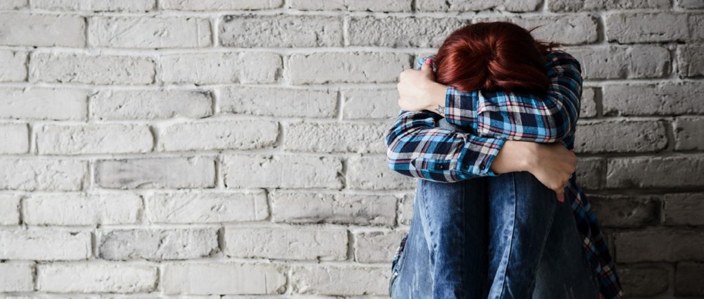 woman leaning against gray brick wall, head down, arms folded over knews and face