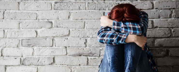 Young adult sitting against brick wall, arms folded over knees, with head bent.