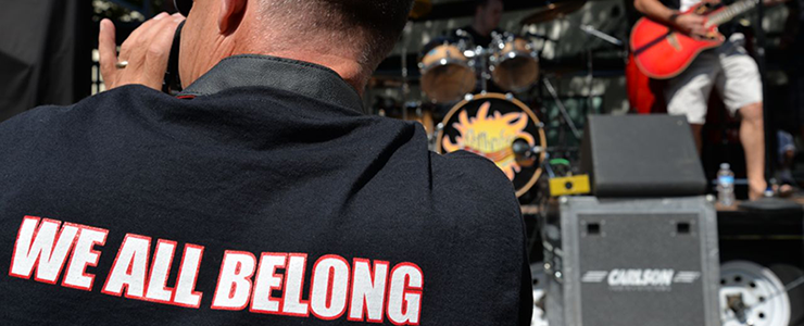 outdoor band playing, back of t-shirt of audience member reads: We All Belong