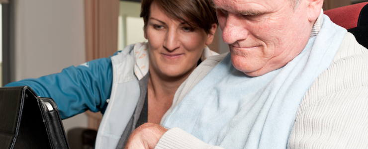 Man in a wheelchair using a laptop with assistance from a care worker.