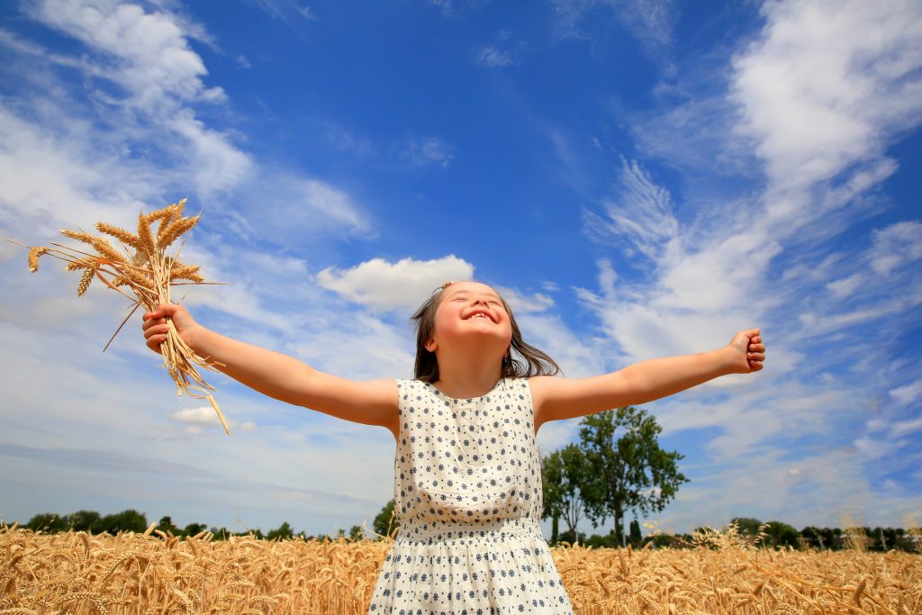 Smiling young girl with Down syndrome in a wheat field with arms outstretched. Sunny, blue skies with fluffy white clouds in background.