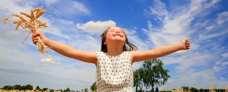 Smiling young girl with Down syndrome in a wheat field with arms outstretched. Sunny, blue skies with fluffy white clouds in background.