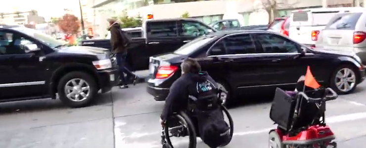 two persons in wheelchairs trying to cross an intersection that's blocked with vehicles in the crosswalk.
