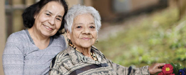 Adult daughter and mother smiling. Blurred outside setting in background.