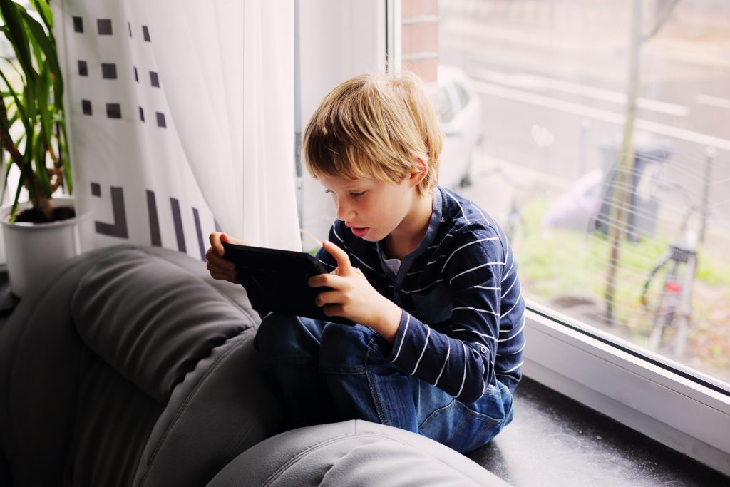 7 year old boy sitting near the window and playing tablet