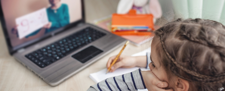 A young girl writing on a piece of paper while watching a teacher on her laptop hold up a math equation.