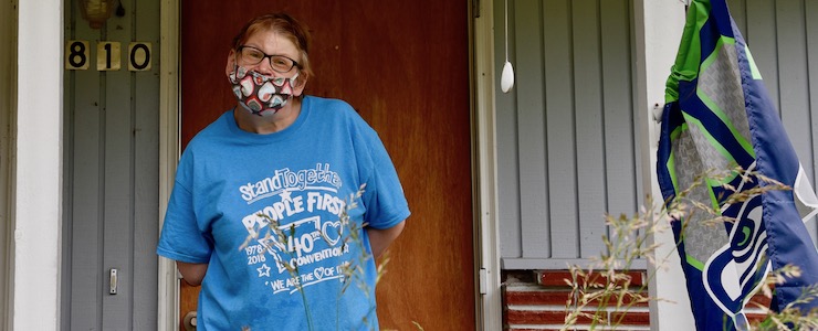 Disabled woman with People First t-shirt wearing a mask, standing outside on a porch.